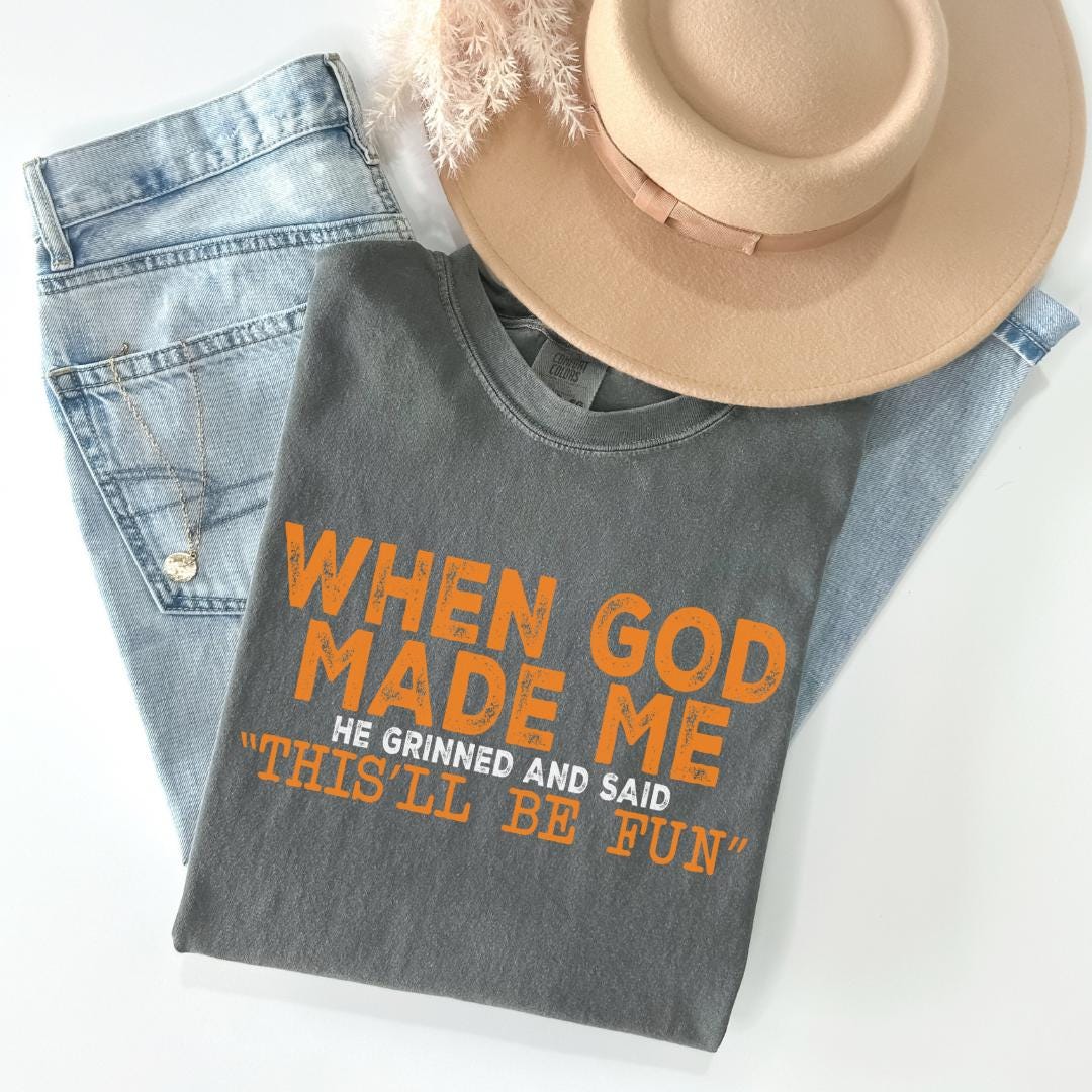 A gray t-shirt with the text "WHEN GOD MADE ME HE GRINNED AND SAID THIS 'LL BE FUN'" printed on it, along with a beige hat and a pair of blue jeans.