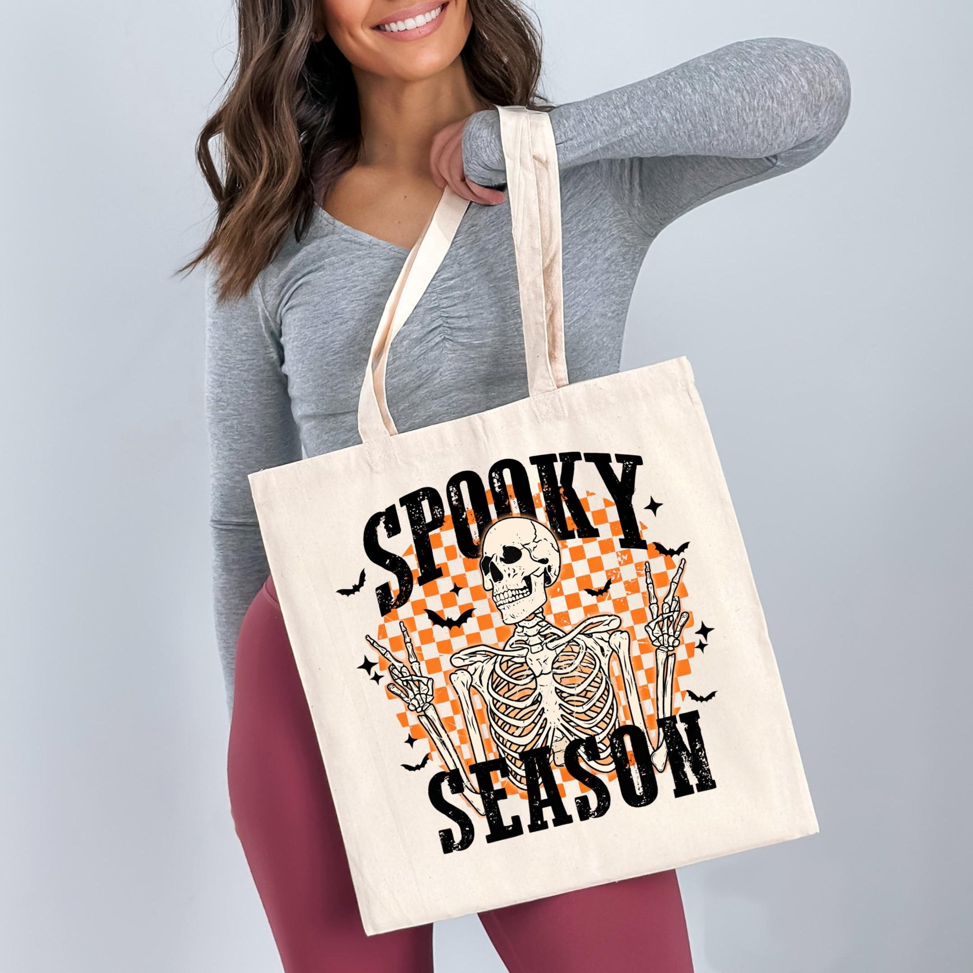 A woman is holding a white tote bag with a Halloween-themed design, including a skeleton and bats, and the text "SPOOKY SEASON".