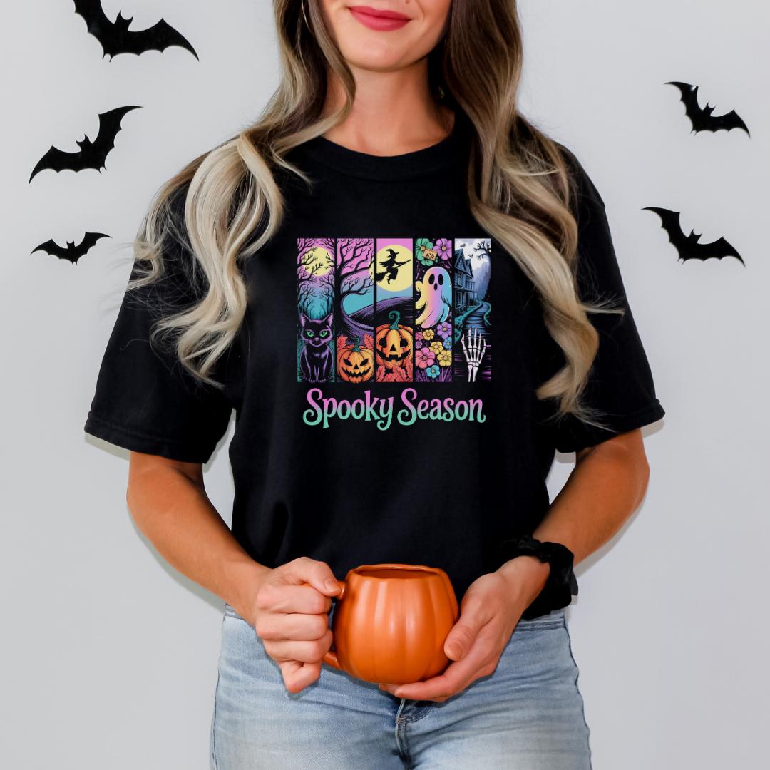 A woman with long blonde hair is holding a pumpkin-shaped mug and standing in front of a wall with bats. She is wearing a black t-shirt with a graphic design featuring various Halloween-themed illustrations.