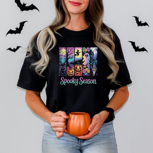 A woman with long blonde hair is holding a pumpkin-shaped mug and standing in front of a wall with bats. She is wearing a black t-shirt with a graphic design featuring various Halloween-themed illustrations.