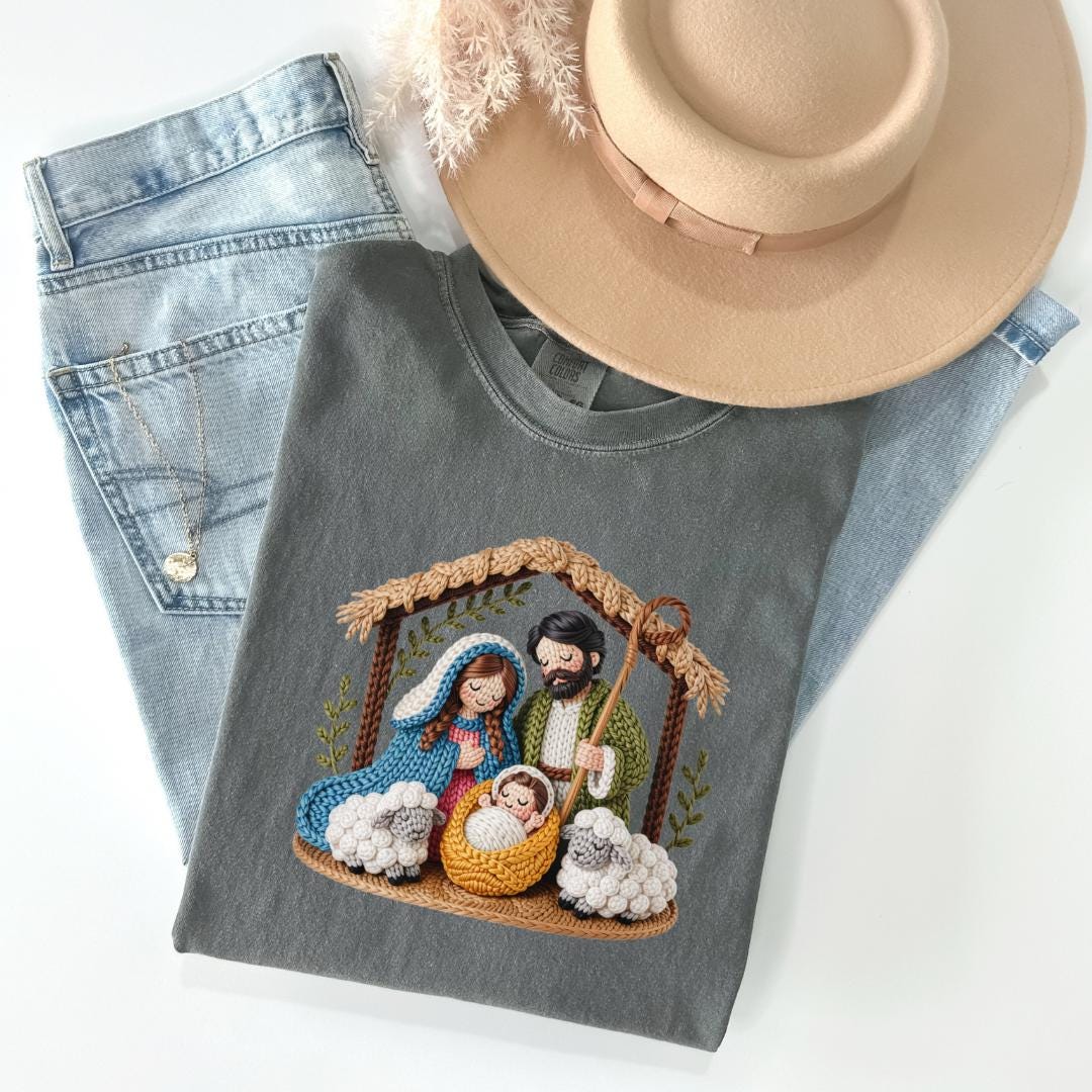 A gray t-shirt with a crochet nativity scene design, a pair of blue jeans, and a beige hat are arranged on a white background.