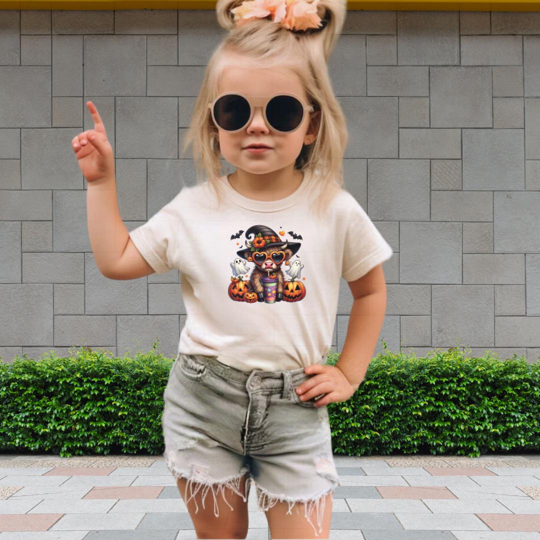 A young girl wearing a white t-shirt with a Halloween-themed graphic, sunglasses, and denim shorts is standing in front of a brick wall.